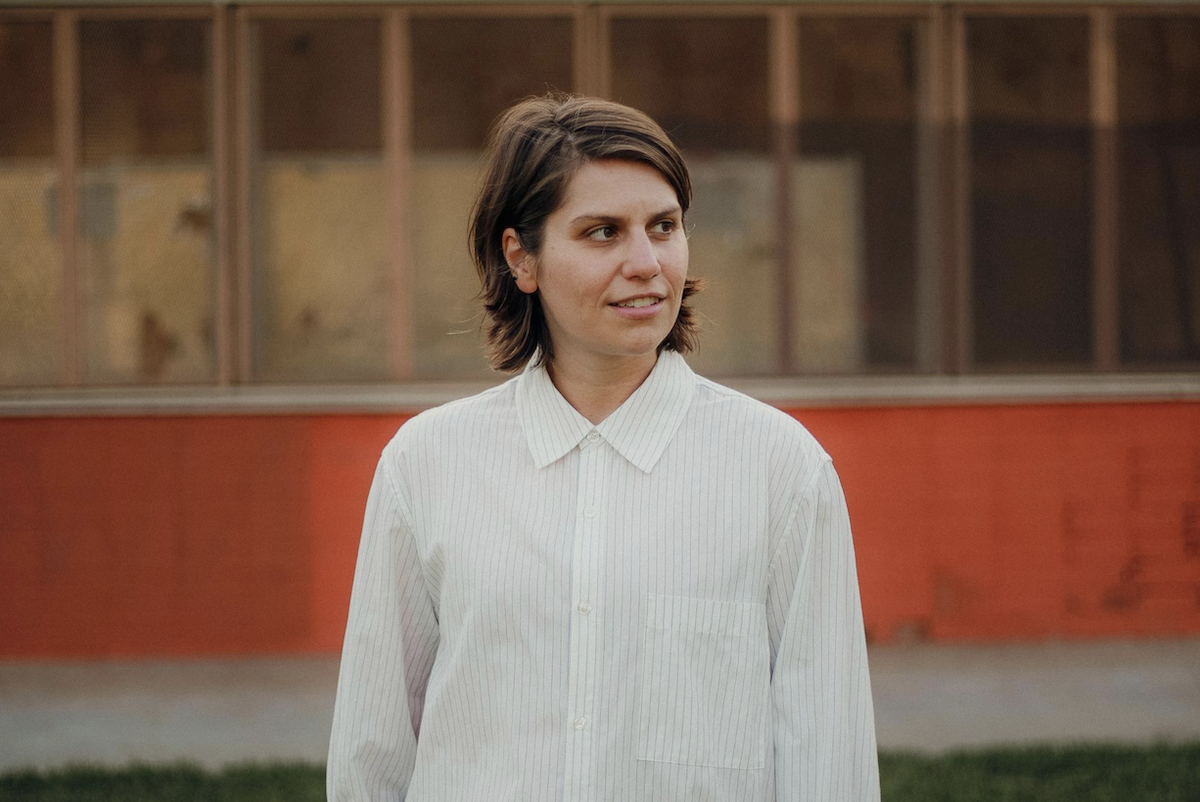 Australian musician Alex Lahey wearing a white pinstripe collared shirt outdoors, looking off-camera against a red wall.